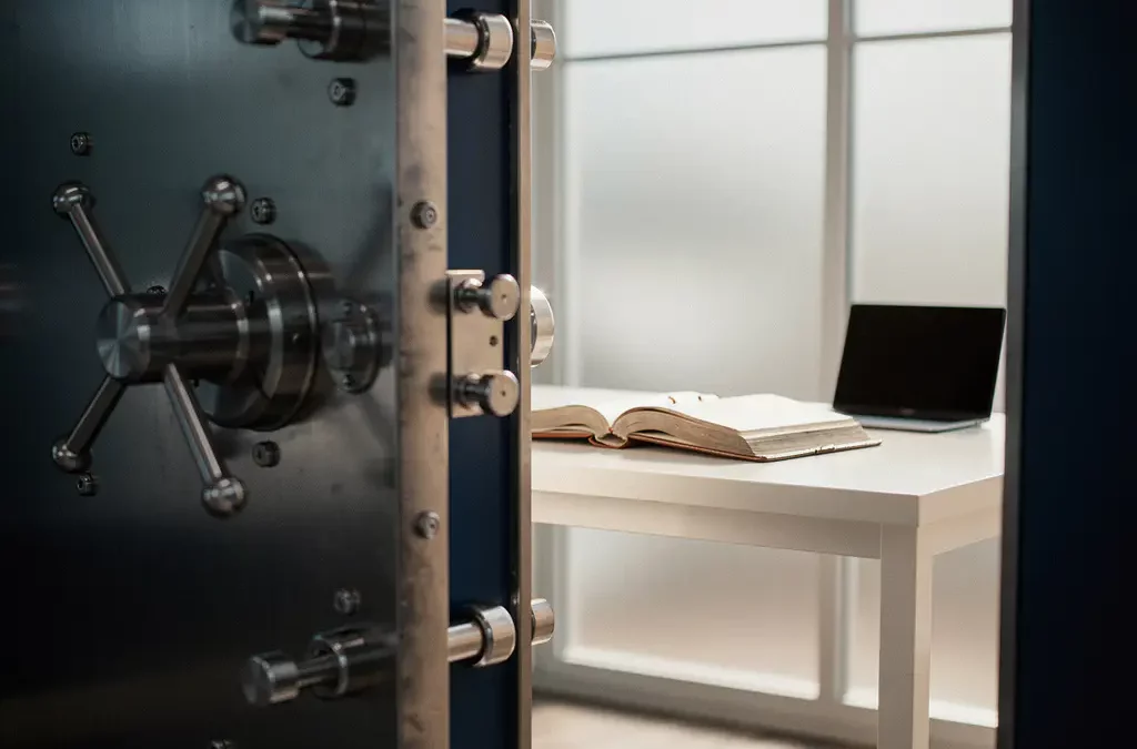 Stalen kluisdeur op een kier met zichtbaar boekhoudwerkplek, laptop en leren grootboek op bureau, zachte kantoorverlichting.